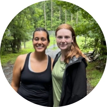 Alia and Amanda, two women, smile at the camera while on a gravel road against a backdrop of green deciduous trees.